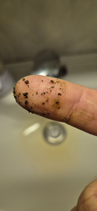 Finger täckt med små, mörka partiklar, förmodligen sediment från en kran, mot en suddig bakgrund av ett handfat.