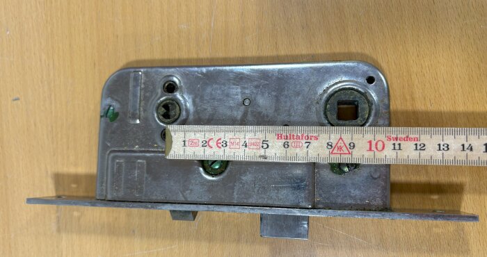 Gammalt dörrlås med centrum-centrum-avstånd på 92 mm, mätt med linjal, ligger på träyta.