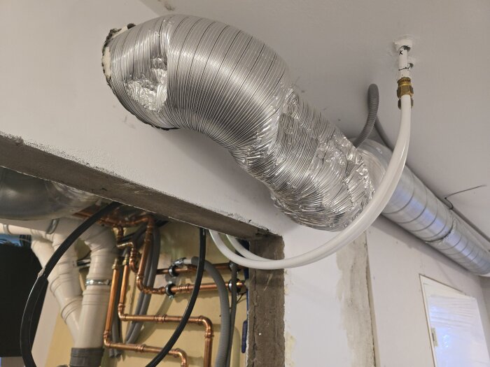 En stålfoliebeklätt ventilationsrör och ett vitt pex-rör i ett maskinrum under renovering, med synliga kopparrör och väggöppningar.