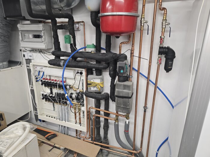 Pex-rör, kopparrör och ett svart magnetitfilter i ett maskinrum. Rör för tilluft, vatten och nätverk syns i fördelarskåpet.