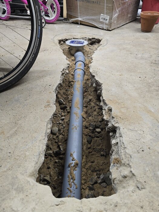 Plaströr i golvets utgrävda schakt, omgiven av makadam, förberett för cement. Bakgrunden visar cykel och kartonger.