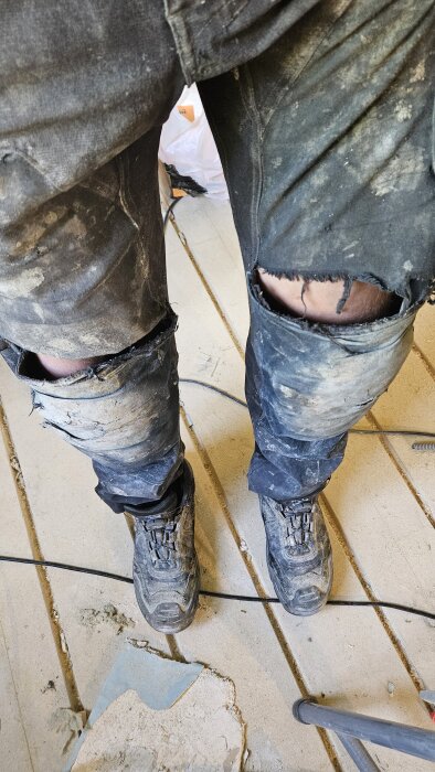 Slitna arbetsbyxor med revor vid knäna och smutsiga arbetsstövlar på ett trägolv under ett byggprojekt.