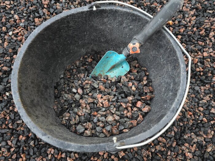 Hink fylld med svart och röd makadam med en blå spade, stående på en yta av makadam.