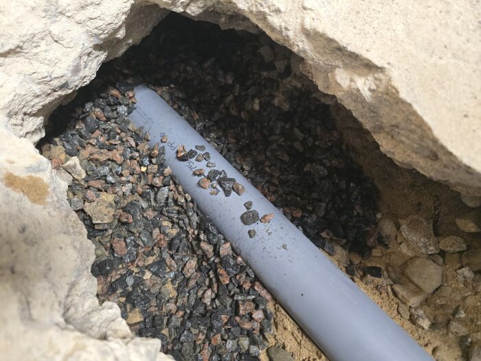 Grått plastvattenrör omgivet av grovt grus, synligt i en öppning i en stengrotta, redo för renovering och betonggjutning.