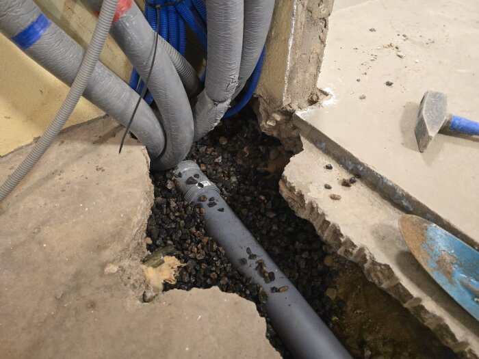 Nedgrävt grått rör omgivet av makadam i ett betonggolv, synliga ledningar och ett blått spadeverktyg intill en upptagen del av golvet.