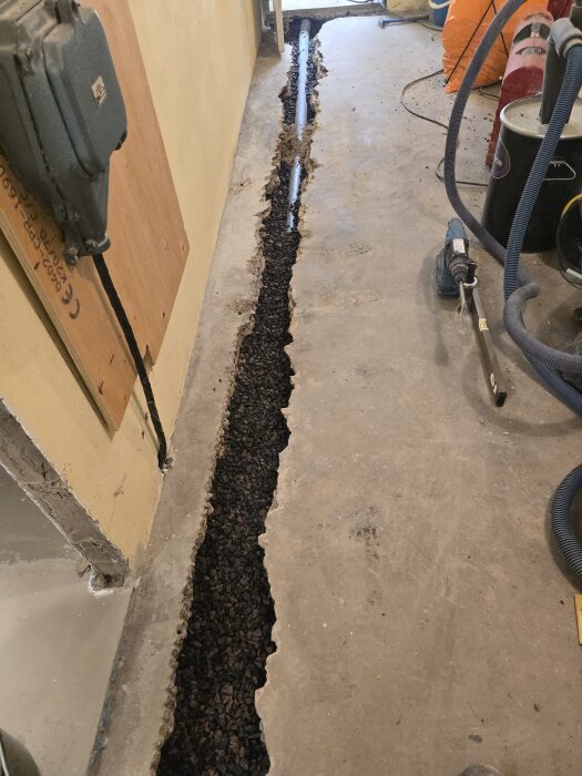Grävd kanal i golvet med synligt avloppsrör, omgiven av fyllnadsmaterial (makadam), verktyg och utrustning runtom.