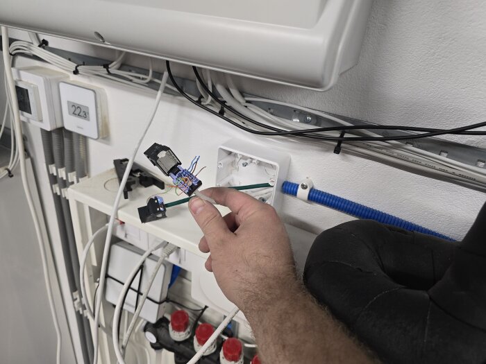 En hand håller en nätverkskontakt med kablar i ett teknikrum, omgivna av olika el- och datorsystem.