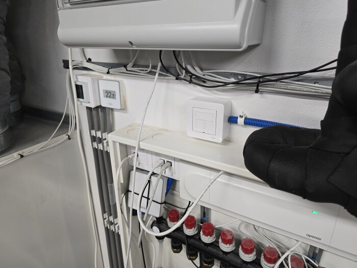 Vägg med rör och kablar i ett maskinrum, termostat visar 22,3°C, nätverkskablar och eluttag synliga, berättelse om renovering och omdragning av rör.
