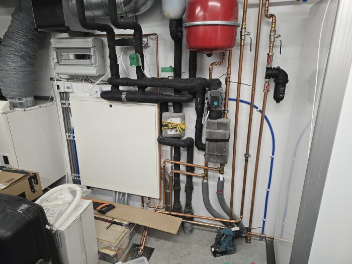 Maskinrum med kopparrörsanläggning, flexibla rör, elektrisk utrustning, och ett svart magnetitfilter monterat på väggen. Rörrenovering pågår.