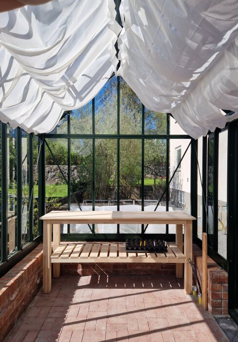 Arbetsbänk i ett växthus med tegelgolv, omgiven av stora fönster och ett tak täckt med vita tygskynken.