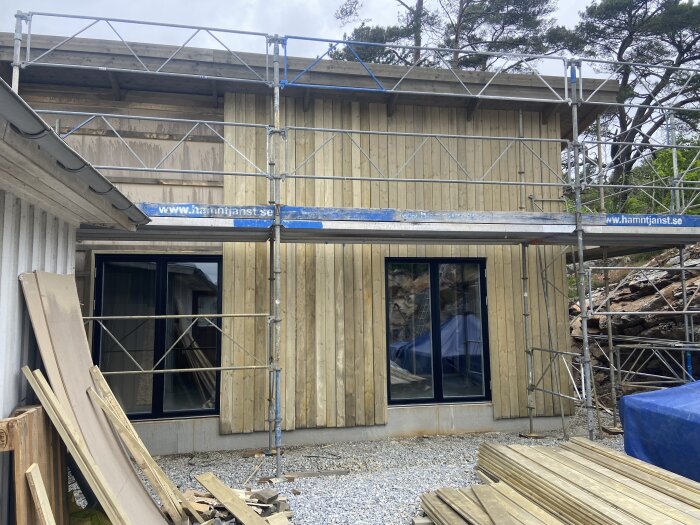 Byggställning runt ett hus med träpanel, visar missfärgning på fasaden ovanför fönster. Byggmaterial ligger på marken framför.