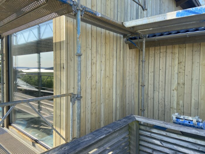 Fasad av träpanel med byggnadsställning, vissa brädor har missfärgningar. Soligt väder och utsikt över landskap syns genom ett stort fönster.