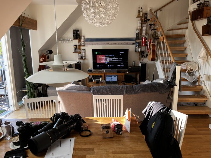 Vardagsrum och kök i öppen planlösning med soffa, TV, kamerautrustning på bordet, trappa till övervåning och dekorativ belysning.