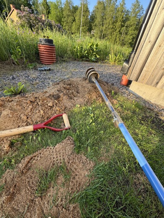 Gräsmatta med ett nytt borrhål, omgivet av jord och verktyg, en grävd grop, metallstång och röd kruka nära ett träplank.