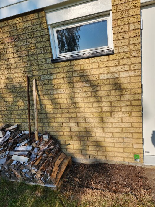 Tegelvägg med fönster och dörr; vedhög och jordyta nedanför. Förberedelse inför installation av robotgräsklippare beskrivs i forumdiskussion.