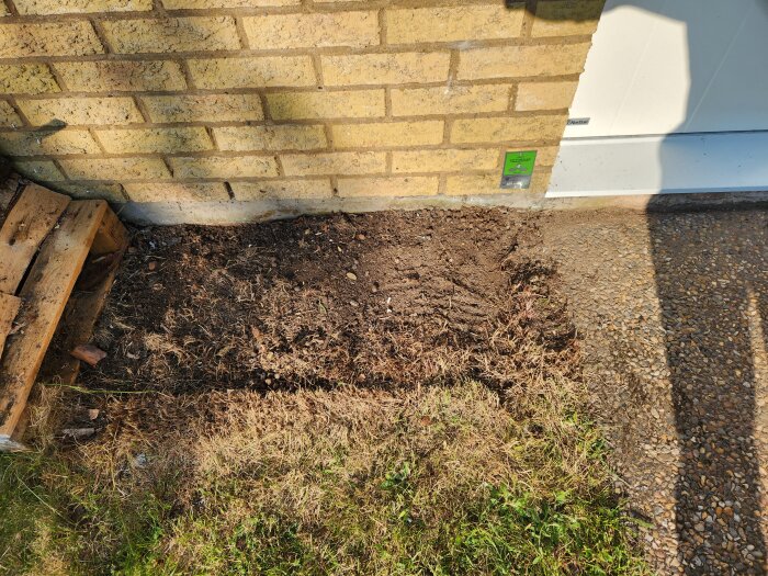 Grusad yta och jord bredvid husvägg. Palldel synlig till vänster. Skuggor på marken. Möjligen förberett för robotgräsklippare.