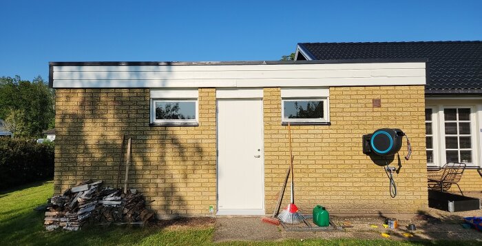 Förrådsbyggnad med gul fasad och vita dörrar, omgiven av vedhögar och trädgårdsredskap. En vattenslang är monterad på väggen.