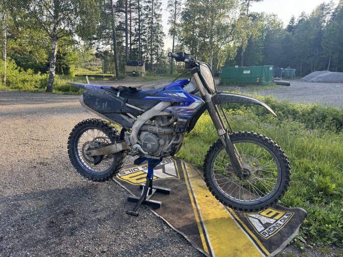 Blå crossmotorcykel stående på en gul och svart matta i en grusig omgivning med gröna träd i bakgrunden.