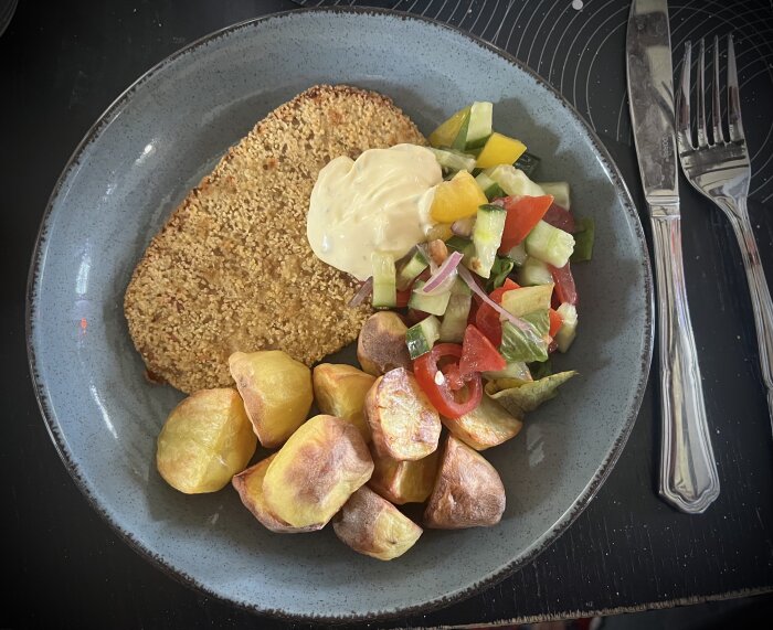 Tallrik med schnitzel, ugnsstekt potatis, sallad och en klick sås, med bestick vid sidan.