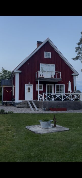 Rött hus med vit dörr och en altan. Trappa till vänster och gräsmatta framför. En pump och ett zinkkar syns på gräsmattan i förgrunden.