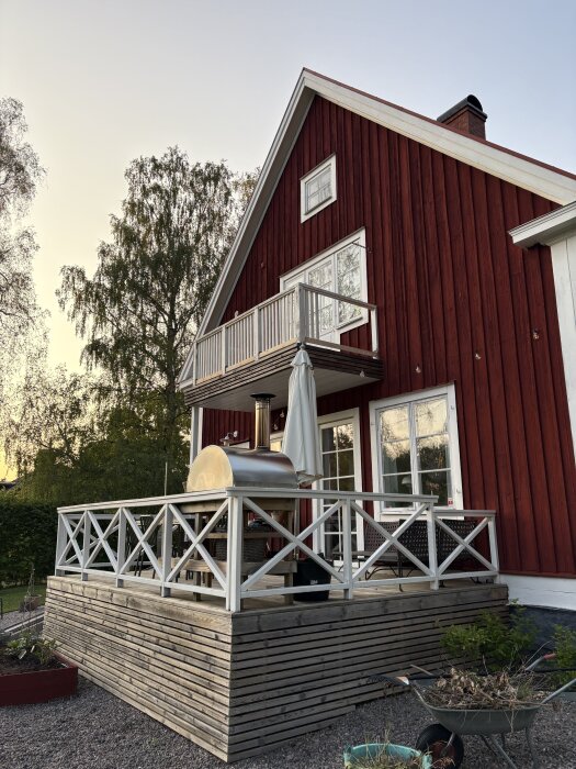 Röd träfasad med en altan försedd med vitt räcke, grill och parasoll. Altanen är byggd ovan mark bredvid gräsmattan.