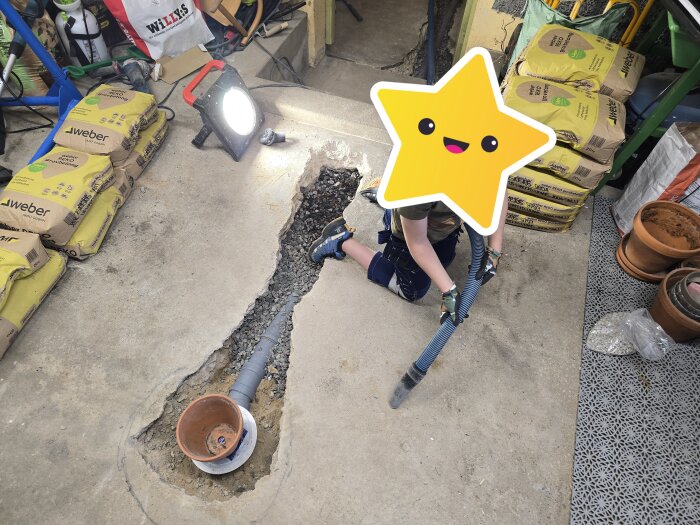 Barn dammsuger runt hål i garagegolv för att förbereda för betonggjutning, omgivna av säckar med betongblandning, under stark bygglampa.