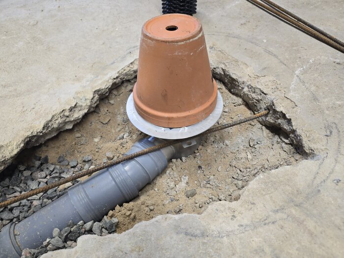 Ett grovhugget betonggolv med en brun golvbrunn täckt av en kruka i ett garage, omgiven av rör och armering.