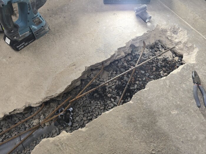 Öppen betongyta i ett garage med synlig armering, grusfyllning och verktyg runtomkring. Ett hål där golvbrunnen kommer installeras.