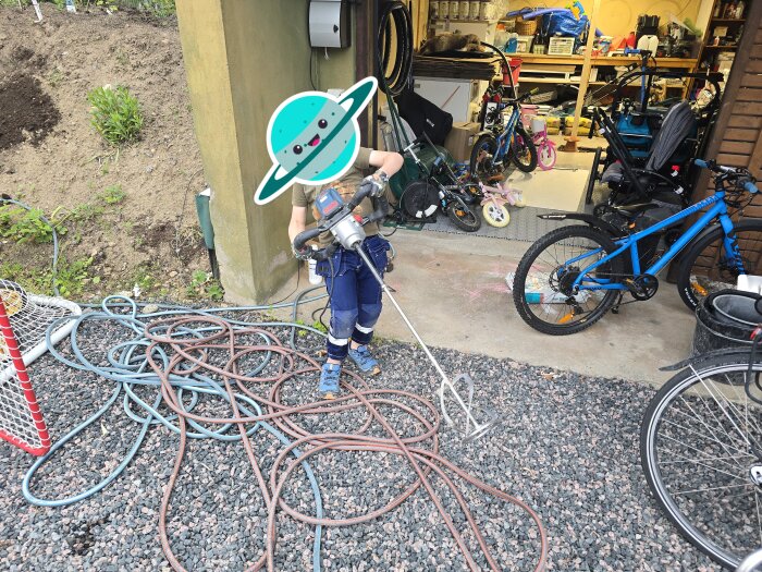 Barn med dräkt och verktyg i handen utanför ett garage, omgiven av cyklar och slangar på marken.