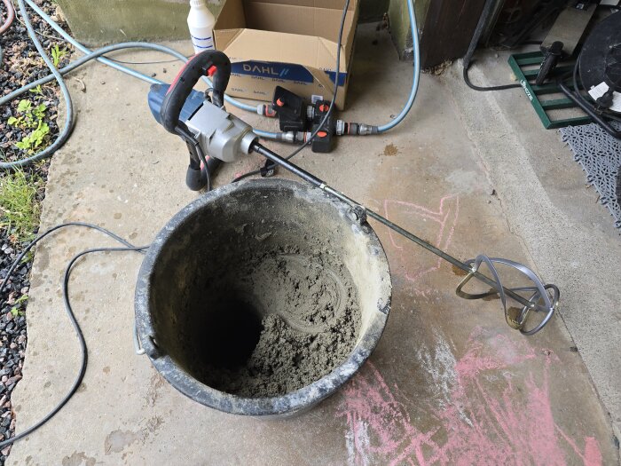 En betongblandare ligger bredvid en hink med blandad betong i ett garage. Verktyg och utrustning syns i bakgrunden.