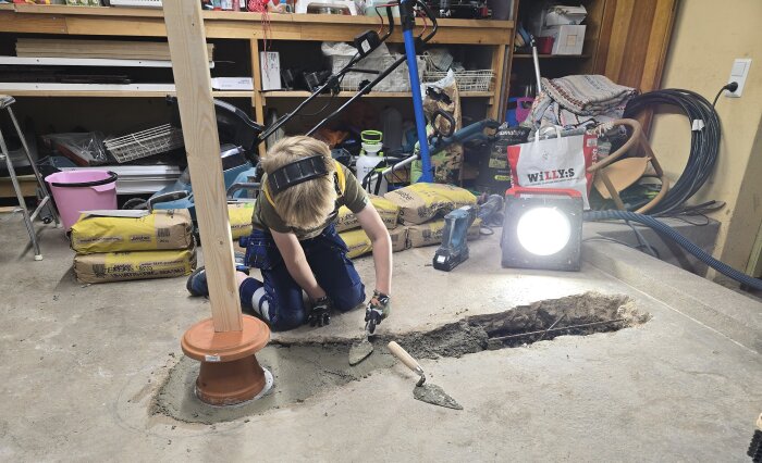 Barn hjälper till med gjutning i ett garage, omgiven av verktyg och byggmaterial. En golvbrunn skyddas av en kruka och en regel.