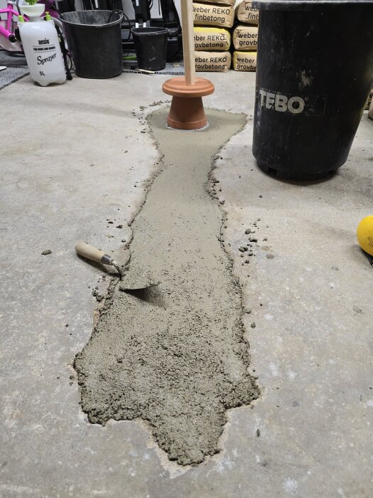 Gjutning av betong i ett garage med en golvbrunn täckt av en kruka och regel ovanpå. Murslev och hink syns bredvid arbetsområdet.