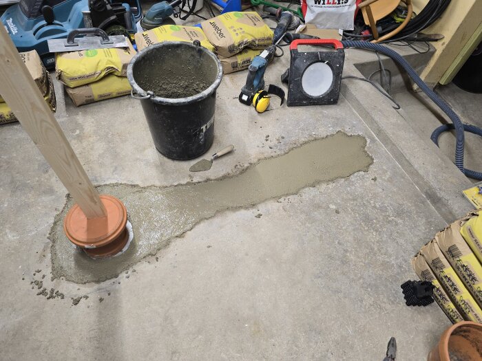 Nyhällt betonggolv i ett garage, omgiven av verktyg och byggmaterial. En golvbrunn skyddas med en kruka och träregel.