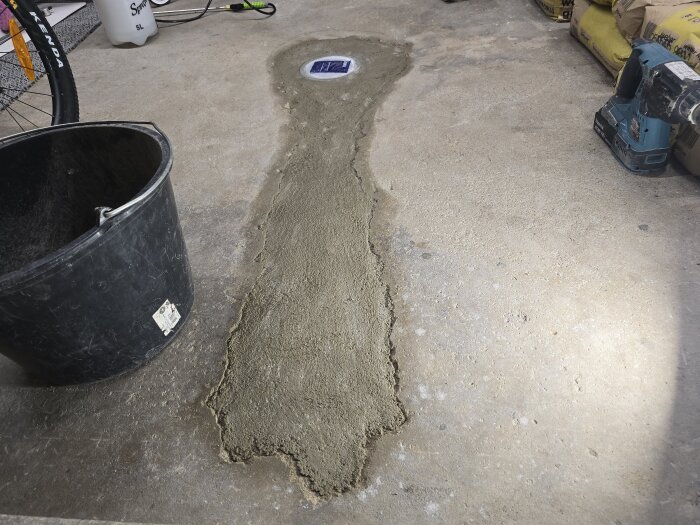 Nybetongad garageyta med synlig golvbrunn, omgiven av verktyg och material; en hink och borrmaskin ses bredvid den nylagda betongen.