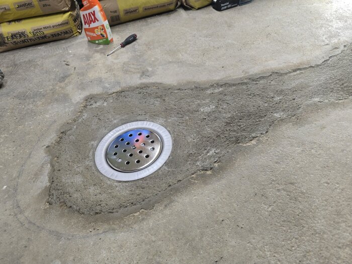 Gjutet betonggolv med nyinstallerad golvbrunn i garage, omgiven av byggmaterial och rengöringsmedel.