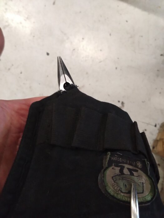 En tång placerad i en svart tånghållare med ett märke synligt på hållaren, fotad mot en ljus bakgrund.