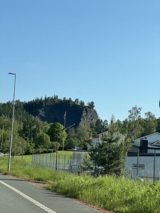 Utsikt över en bergskulle med träd, stängsel och hus i förgrunden under klarblå himmel.