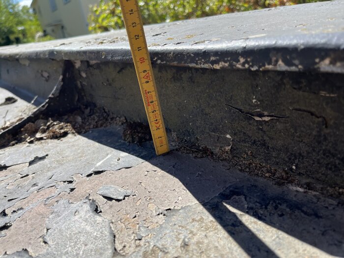 Mätstock på takkant med uppvik, lövansamling och avflagad färg på plåttak.