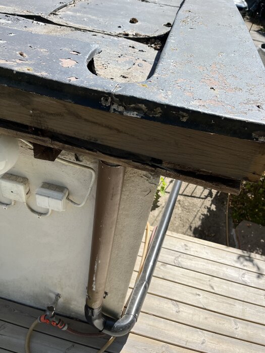 Slitet bandtäckt plåttak med uppvik i bakkant visas på ett garage, saknar hängränna. Avrinningsrör syns vid sidan av byggnaden.