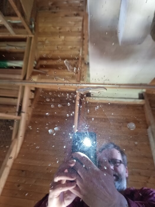 Fotografi av en person i en vind med träpaneler, som speglas i en våt yta med vattenstänk.