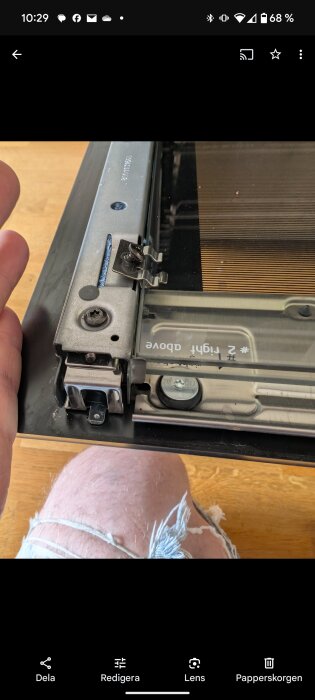 Svart metallbeslag med skruvar och klämmor på glasram, omgiven av träyta och hand i jeans.