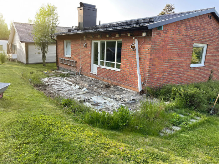 Tomt vid tegelhus med markarbete för ny altan, täckt med plastduk. Huset har brun tegelfasad och omgivande gräsmatta.