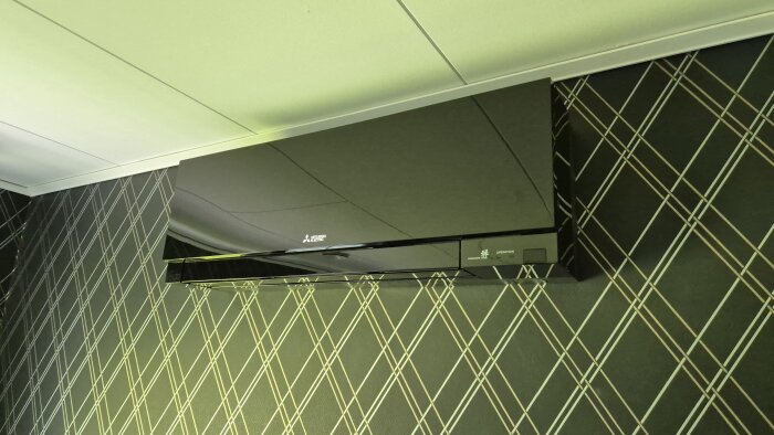 Svart väggmonterad luftvärmepump från Mitsubishi Electric på mönstrad tapet, installerad för att ge kyla med låg ljudnivå i hemmet.