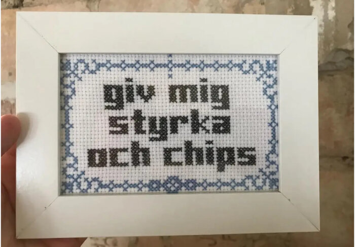 Broderad tavla i vit ram med texten "giv mig styrka och chips" omgiven av blått mönster.