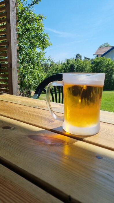 En kall öl i ett frostat glas står på ett träbord utomhus med gröna träd och sommarhimmel i bakgrunden.