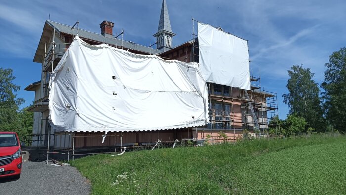 Hus inlindat i byggställningar och vita presenningar, med en kyrkliknande spira i bakgrunden och en röd bil framme till vänster.