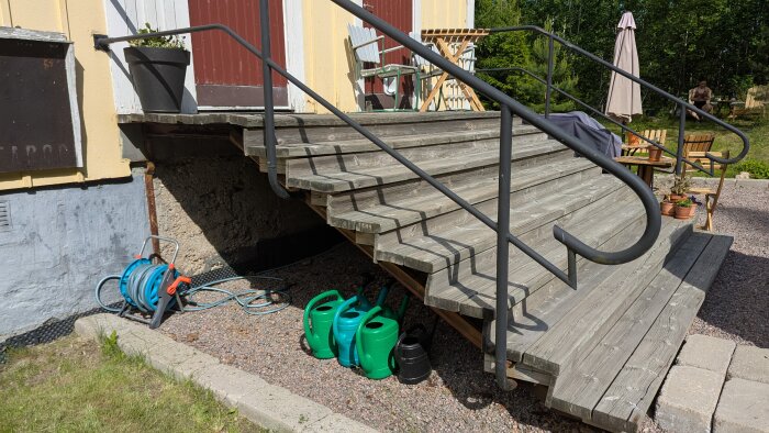 Trätrappa med svart räcke fäst vid gul fasad. Vattenkannor och vattenslang syns under trappan. Trädgårdsmöbler och parasoll i bakgrunden.
