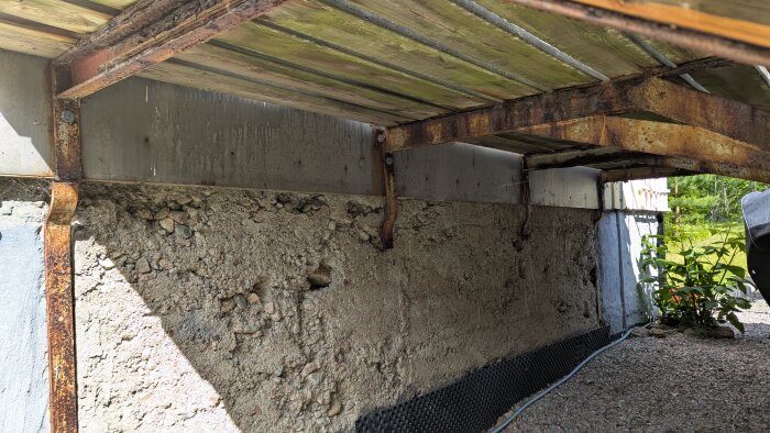 Rostig stålkonstruktion fäst i en betongvägg, med träbalkar ovanpå och grus samt vegetation i bakgrunden.