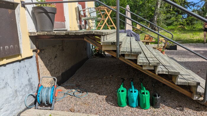 Utomhustrappa av trä med metallräcke, planteringskärl vid dörren, vattenkannor och trädgårdsslang under trappan, gårdsmiljö i bakgrunden.
