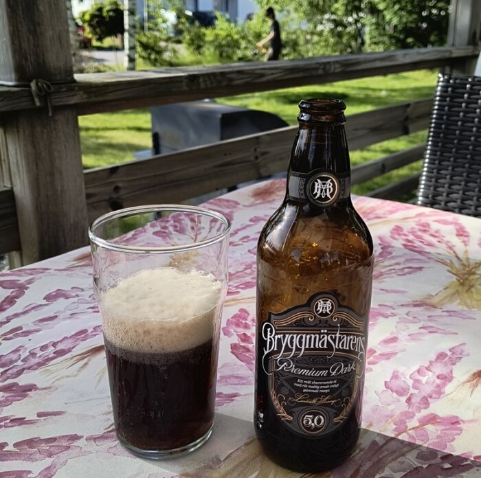 Flaska och glas med öl på ett utebord, person i bakgrunden med gräsklippare, grill synlig.